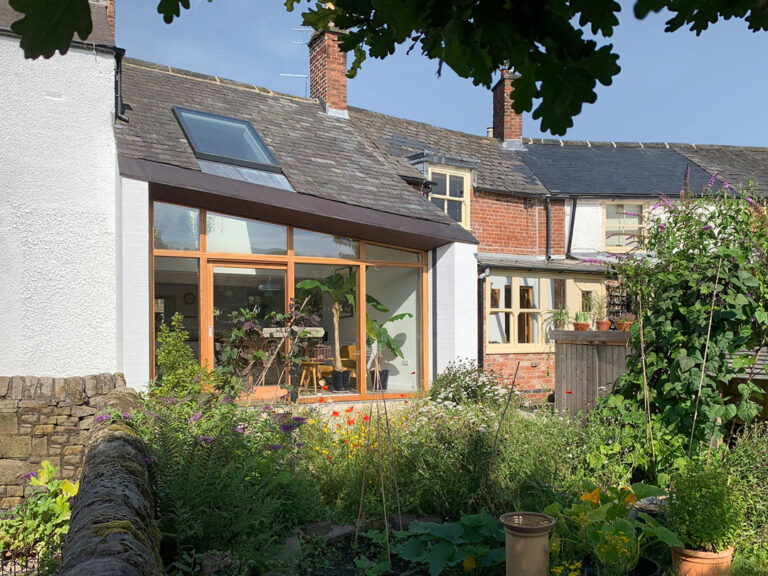 A charming cottage with a modern glass extension and skylight, surrounded by greenery and colorful flowers. The garden features a variety of plants and a stone wall in the foreground. Brick and white exterior with chimney stacks complete the scene.