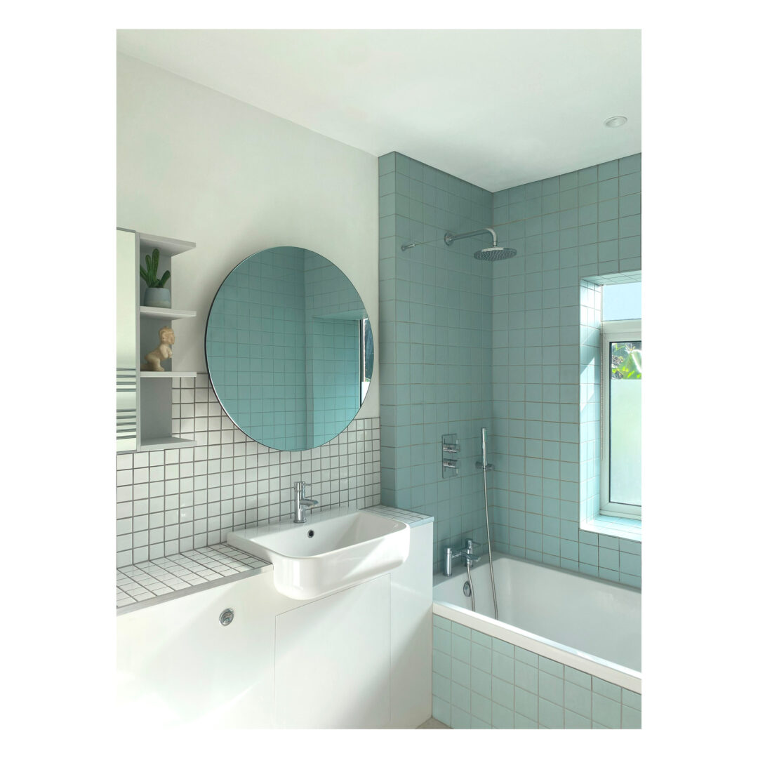 Modern bathroom with light blue square tiles on the walls. A round mirror hangs above a white sink. Theres a bathtub with a showerhead, and a small window lets in natural light. Minimalist shelving is visible to the left.