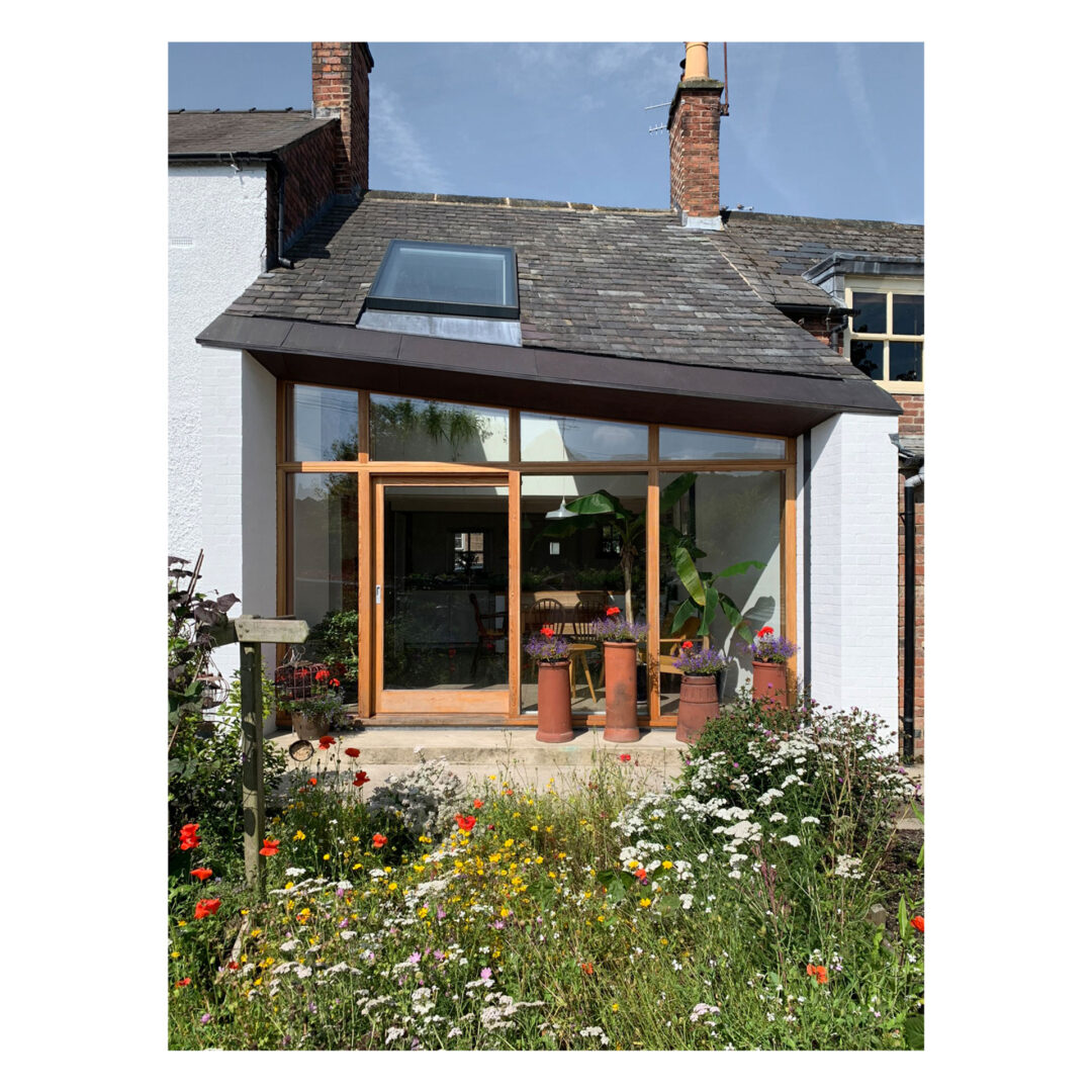 A charming house with a sloped roof, large glass windows, and lush greenery. The entrance is surrounded by colorful wildflowers and clay pots. A cozy interior with visible plants inside complements the natural garden below.