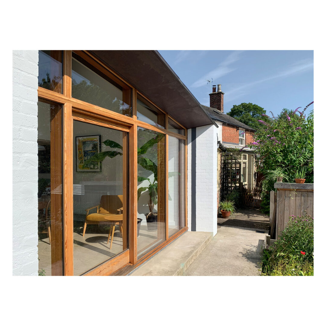 A modern house exterior designed by renowned Bristol architects features large wooden-framed windows reflecting sunlight. Inside, a mid-century chair is visible, surrounded by plants. The pathway leads to a garden with lush greenery and another brick house in the background under a clear blue sky.