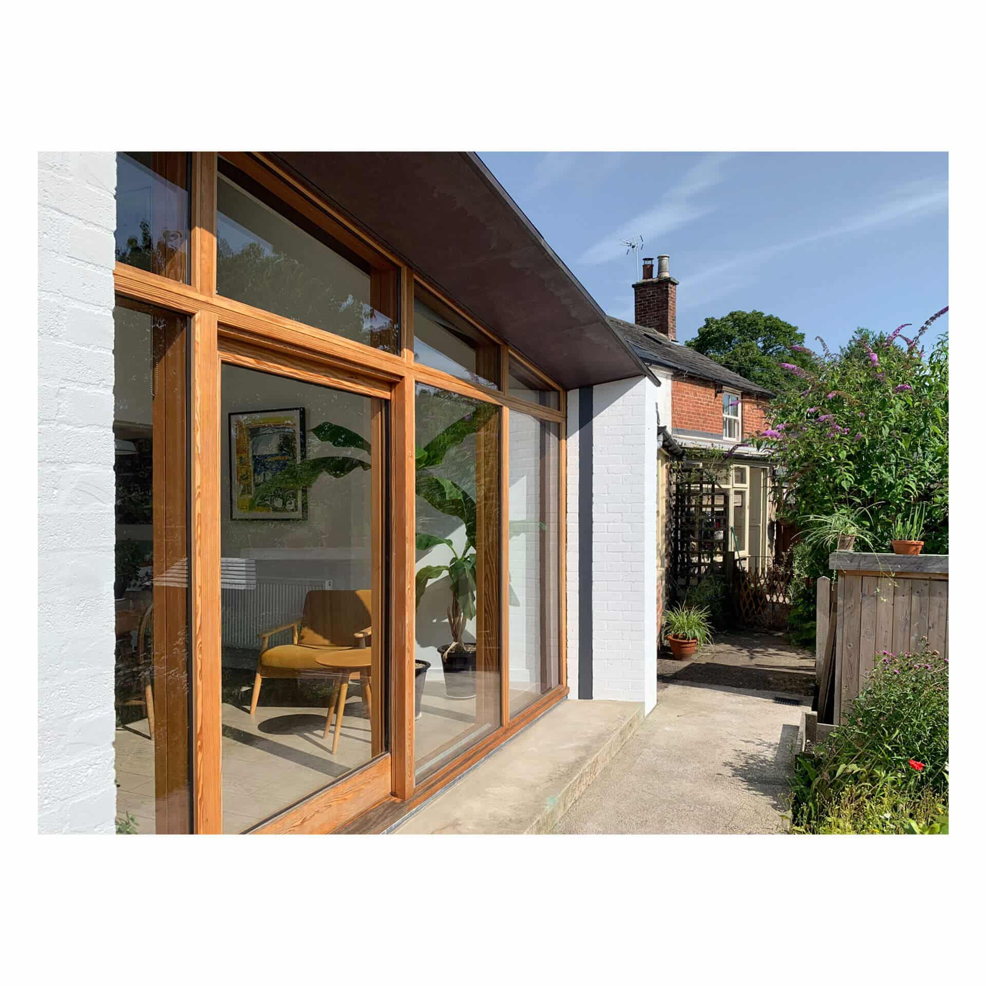 A modern house exterior designed by renowned Bristol architects features large wooden-framed windows reflecting sunlight. Inside, a mid-century chair is visible, surrounded by plants. The pathway leads to a garden with lush greenery and another brick house in the background under a clear blue sky.
