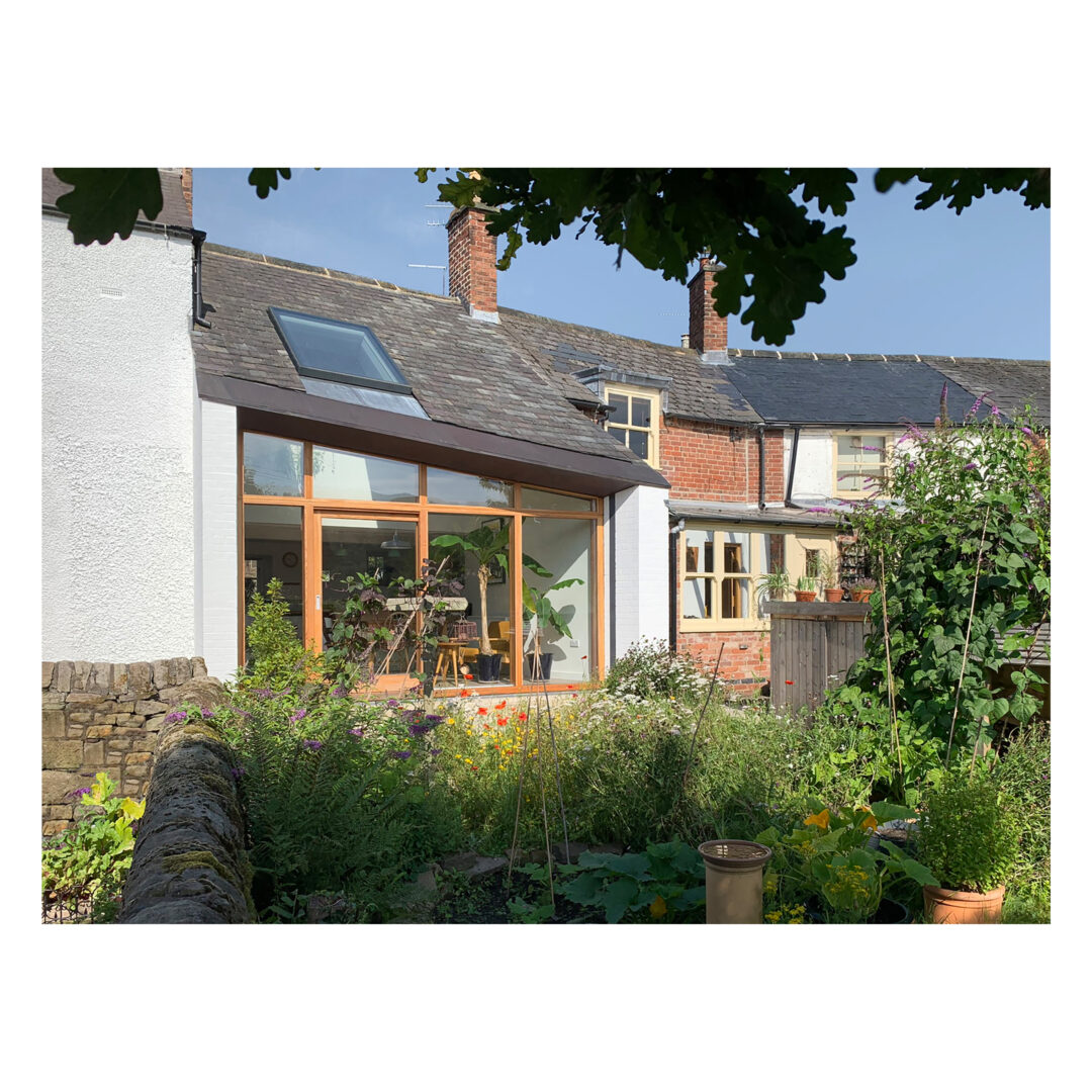 A quaint house with a mix of brick and white walls, featuring a glass extension with wooden frames. A lush garden with various plants and flowers occupies the foreground, partially shaded by tree branches.