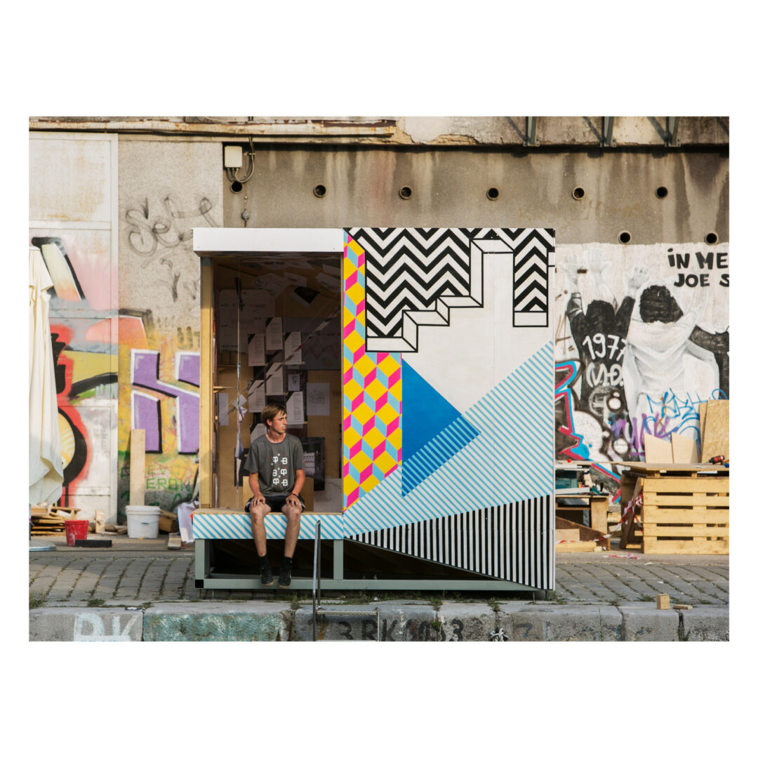 A man sits on a small, colorful structure with geometric patterns and graffiti-style art. The background features a wall covered in various graffiti designs. He wears a casual outfit and a hat, seated on the edge of the structure.