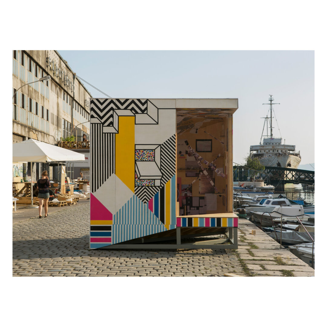 A colorful geometric art installation stands on a cobblestone dock near boats and a ship. The structure, featuring vibrant patterns and stripes, is next to a weathered building and people strolling by.