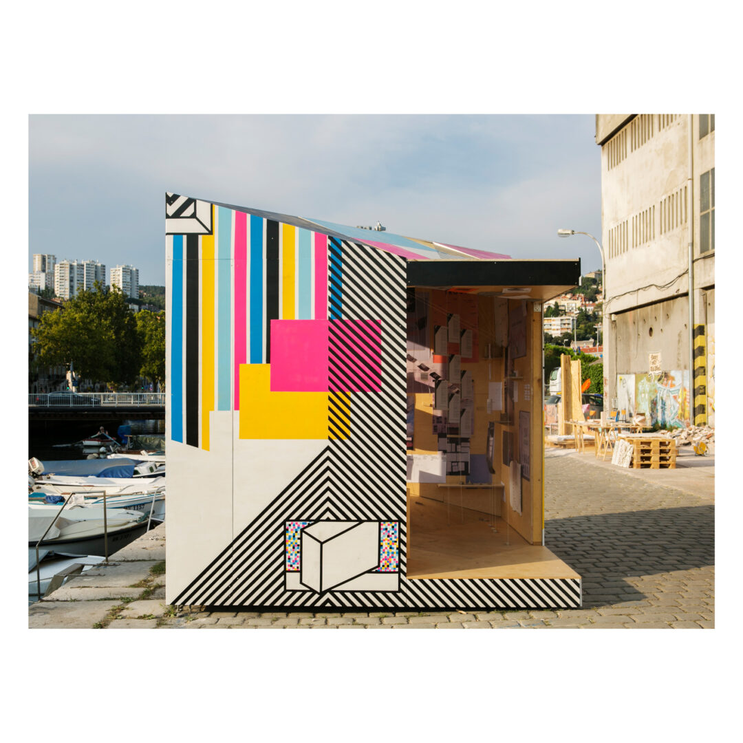A small, modern kiosk with colorful, geometric designs on its exterior stands on a paved walkway near boats and buildings. The open front reveals books and a chair inside. Trees and urban structures are visible in the background.
