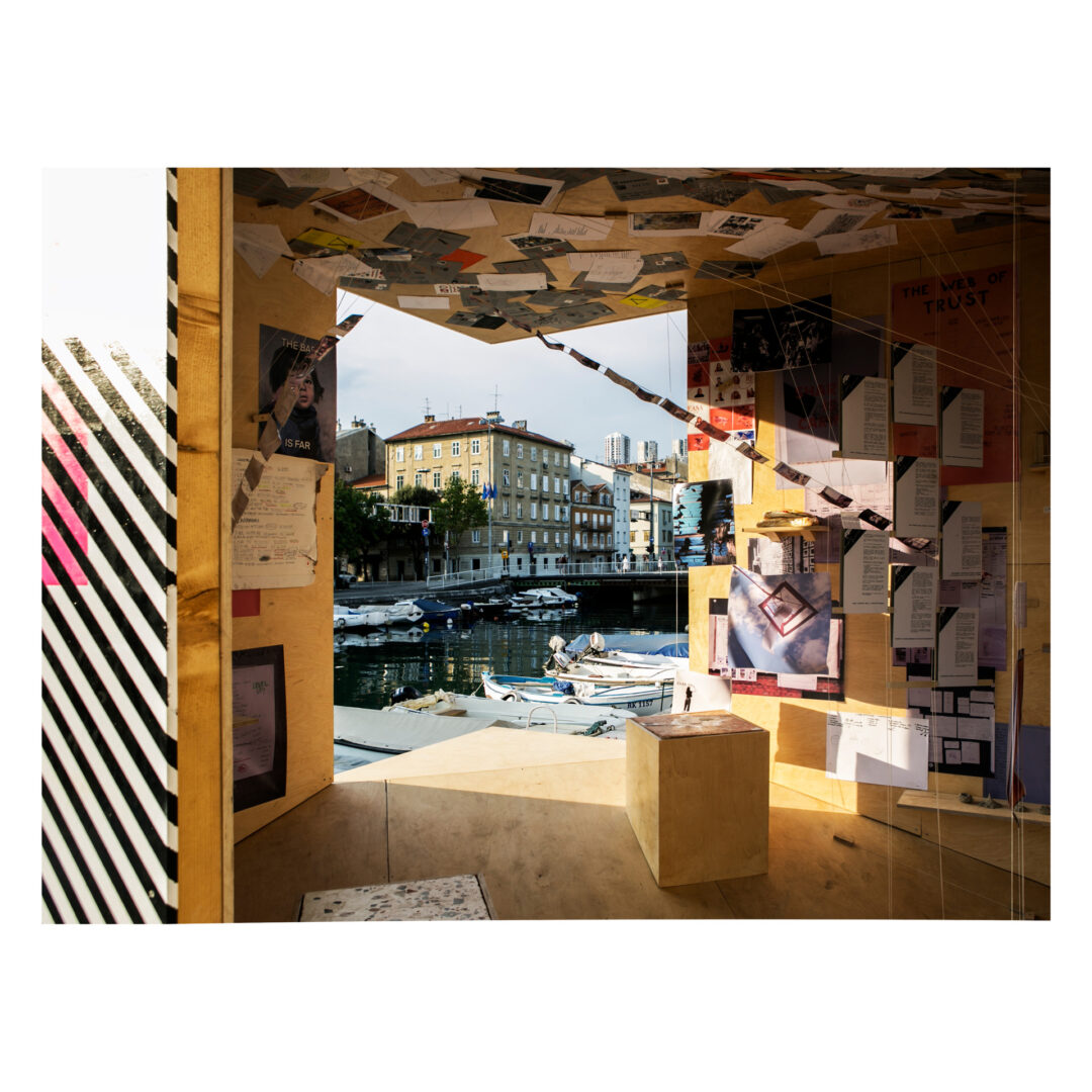 View from inside a wooden, creative installation covered with papers and photos, overlooking a sunlit harbor with boats. A building with a mural is visible across the water, and sunlight casts intricate shadows inside the space.