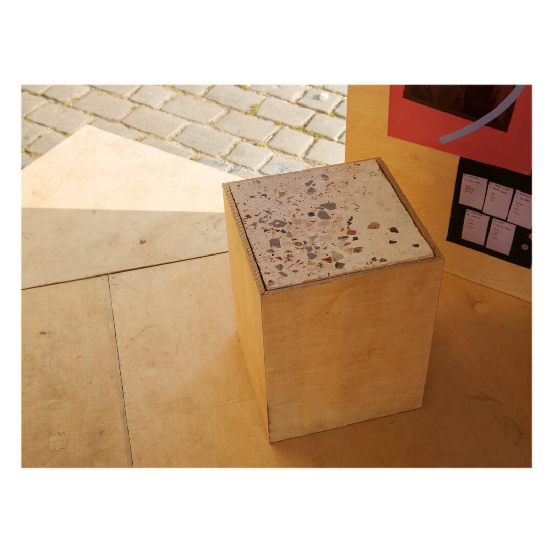 Wooden cube with a terrazzo top placed on a plywood floor, surrounded by walls. A cobblestone pavement is visible outside. A section of a wall on the right features assorted images and cards.