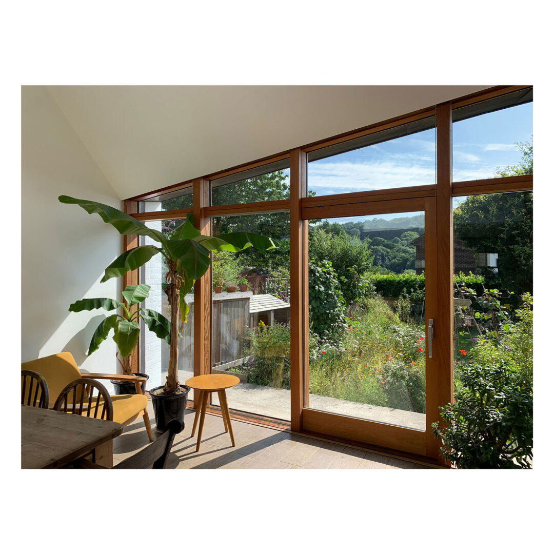 A bright room with large wooden-framed windows overlooking a lush garden. Sunlight streams in, illuminating a banana plant, a wooden stool, and a yellow armchair. The scene is tranquil and surrounded by greenery.