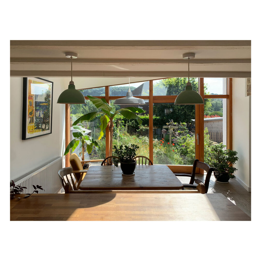 Sunlit dining area with a wooden table and chairs, surrounded by potted plants. Large windows overlook a lush garden. Two green pendant lights hang from the ceiling. Framed art is visible on a white wall to the left.