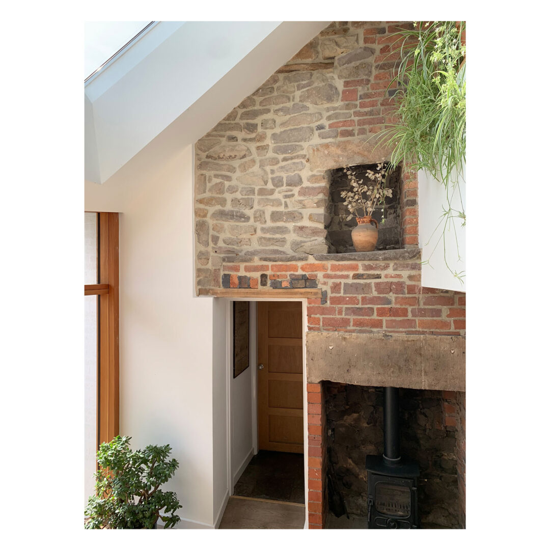 Interior view showcasing a rustic stone and brick wall with a wooden door. Features a small fireplace with a wood-burning stove and a potted plant hanging above. A sloped skylight and a large window brighten the space.
