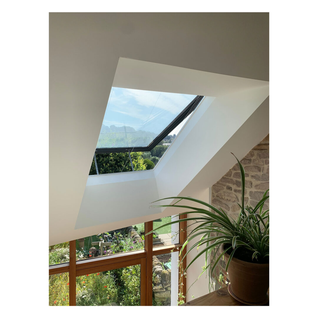A triangular skylight in a room with white walls and a stone accent. Below the skylight, theres a large potted plant on a wooden surface. Outside, lush greenery and a clear blue sky are visible.