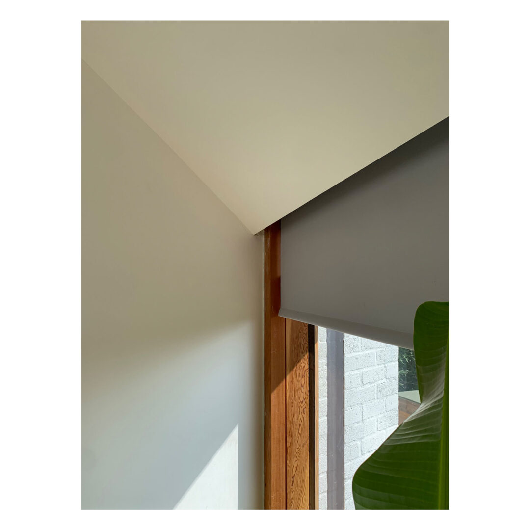 A modern interior corner featuring a slanted ceiling, a wooden window frame, and a partially lowered gray roller blind. Sunlight creates contrasting light and shadows on the white walls, with a green plant leaf visible at the bottom right.