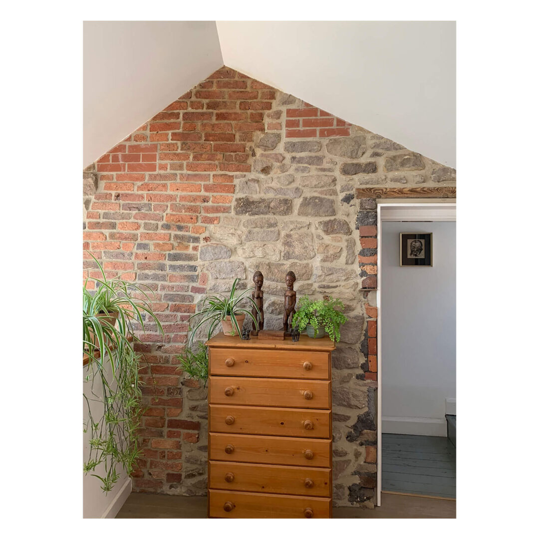 A room with a high sloped ceiling featuring a mix of red brick and stone walls. A wooden chest of drawers is adorned with green plants and two wooden sculptures. An open doorway leads to another space with a framed picture on the wall.