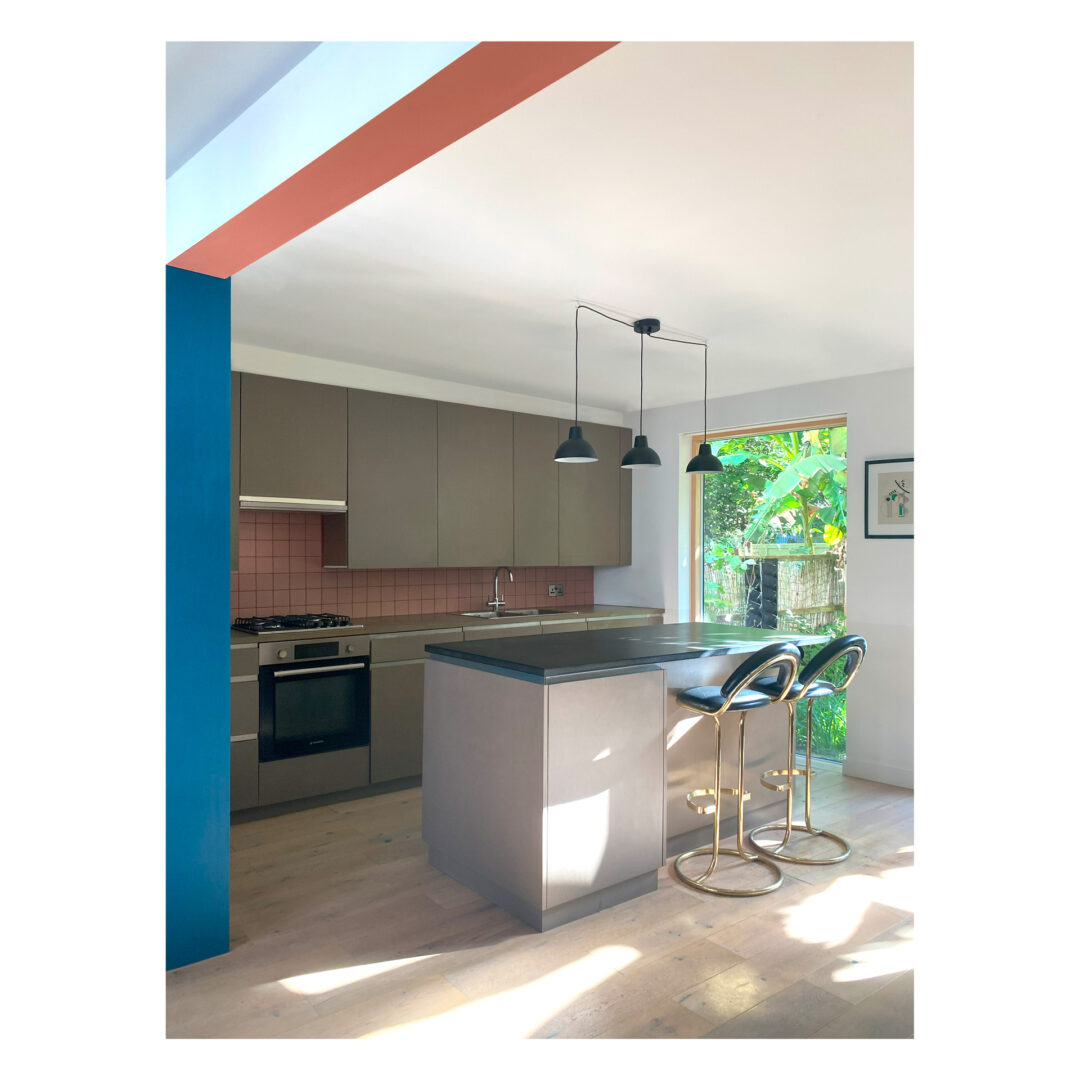 Modern kitchen with grey valchromat cabinets, black countertops, and a pink tile backsplash. Two gold and black bar stools are at the island. Three pendant lights hang above. Large window shows greenery outside. Bright natural light fills the space.
