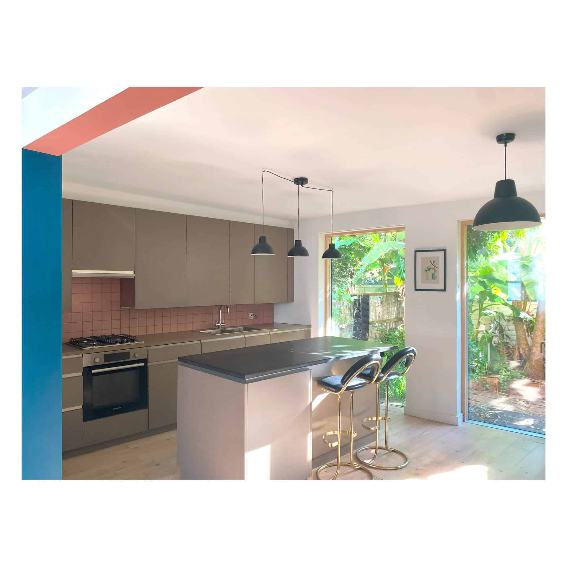 Designed by renowned Bristol architects, this modern kitchen boasts grey cabinets and a large island with black and gold stools. Three pendant lights elegantly hang overhead, while a large window and glass door frame lush greenery outside, bathing the space in natural light.