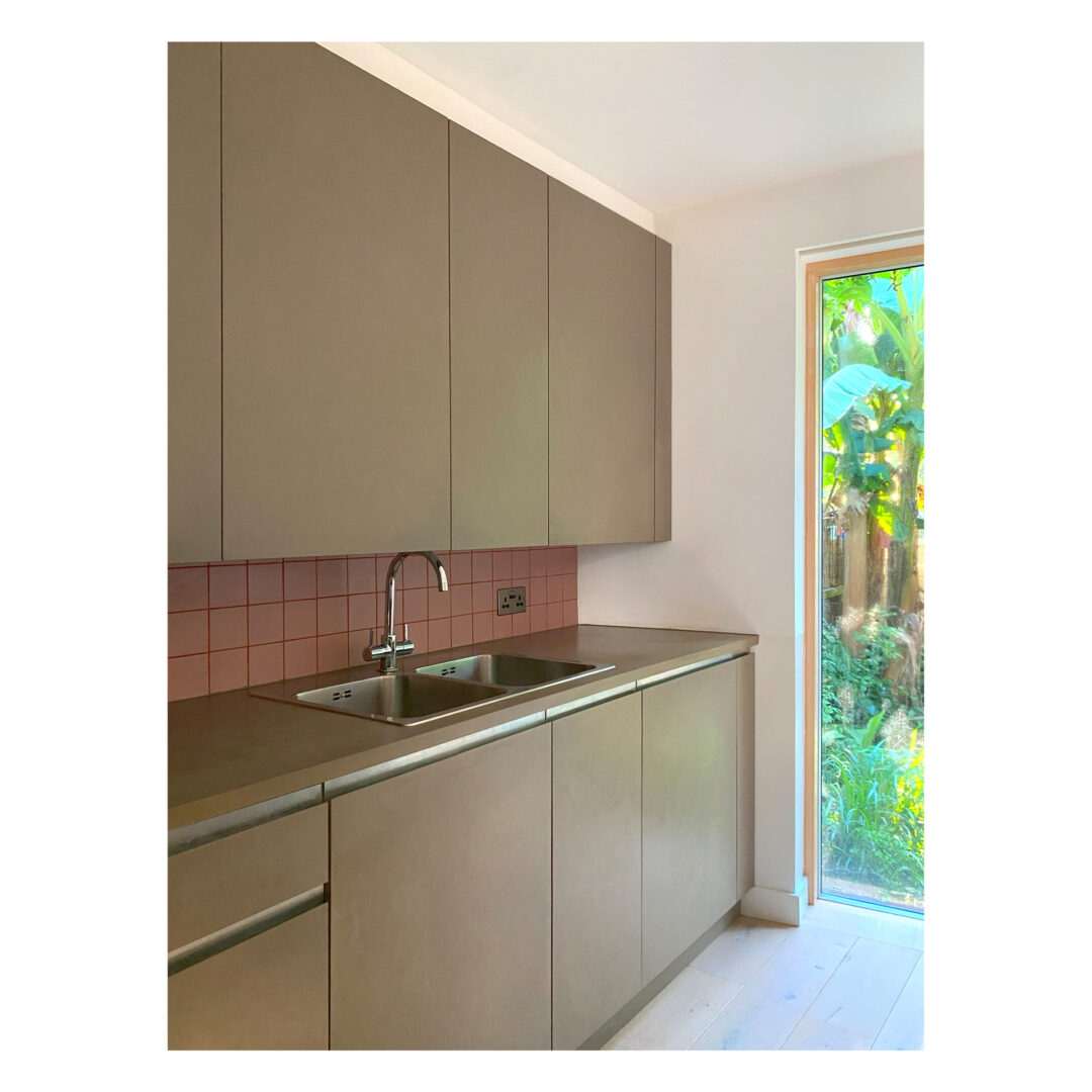 Minimalist kitchen with sleek grey cabinets and a double stainless steel sink. A pink tiled splashback adds contrast. A large window on the right side offers a view of lush green plants outside, allowing natural light to brighten the space.