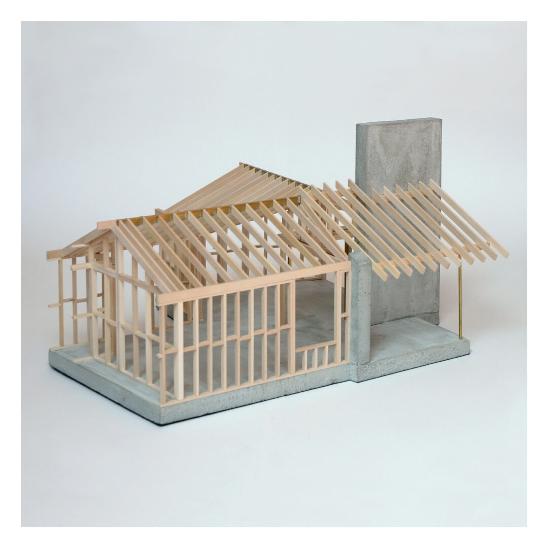 A small architectural model of a wooden house frame on a concrete base, featuring a visible wooden skeleton structure and roof trusses, with a concrete wall section on one side.