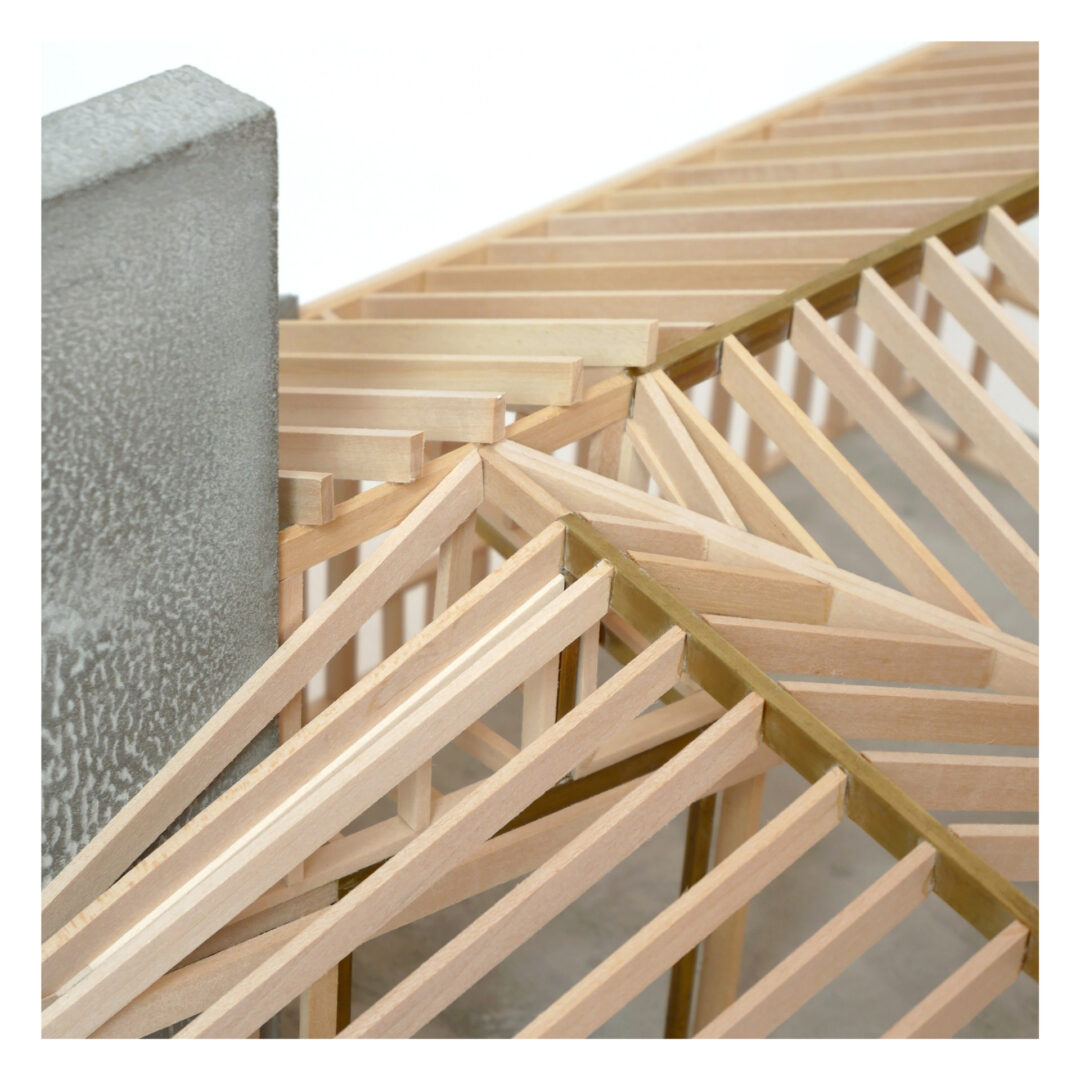 A close-up of a wooden architectural model showcasing an intricate framework of intersecting beams. The beams are arranged in a symmetrical pattern next to a textured gray wall, highlighting structural design elements.