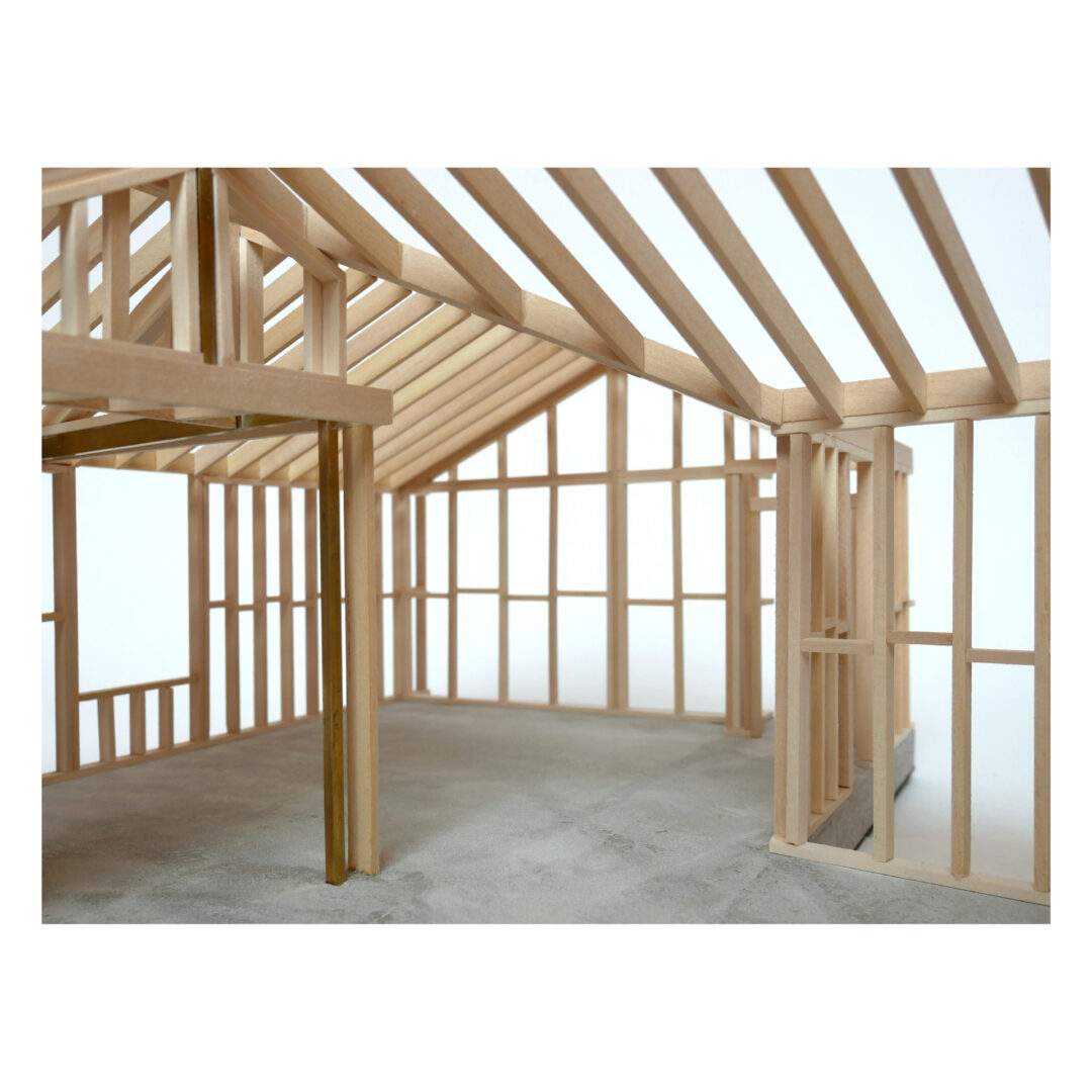 Scale model of a wooden building frame, showing beams and supports on a concrete base. The structure features a framed roof with detailed trusses and open sides, highlighting the construction's skeletal form.