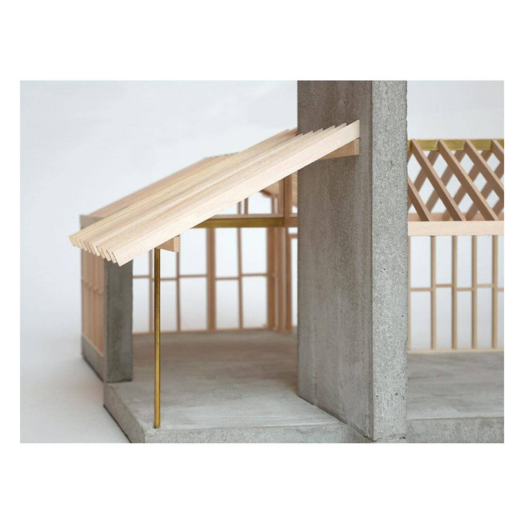 An architectural model with a wooden slatted roof and a brass support pole on a concrete base. The structure shows a partial wall and a grid-like wooden framework. The composition creates a modern, minimalistic aesthetic.