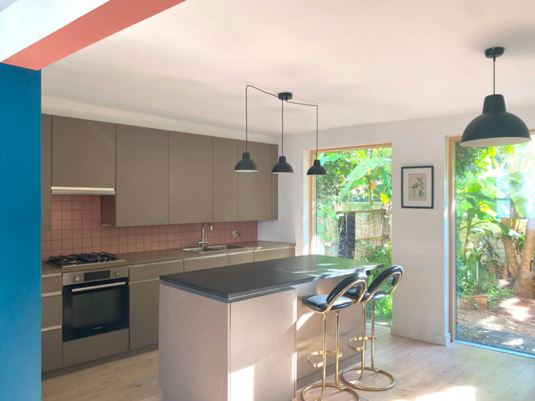 Modern kitchen with brown cabinets, a black countertop island, and three pendant lights. Two chairs are at the island. A large window reveals lush greenery outside, adding natural light to the space—an exquisite feature of a recent house extension in Bristol.