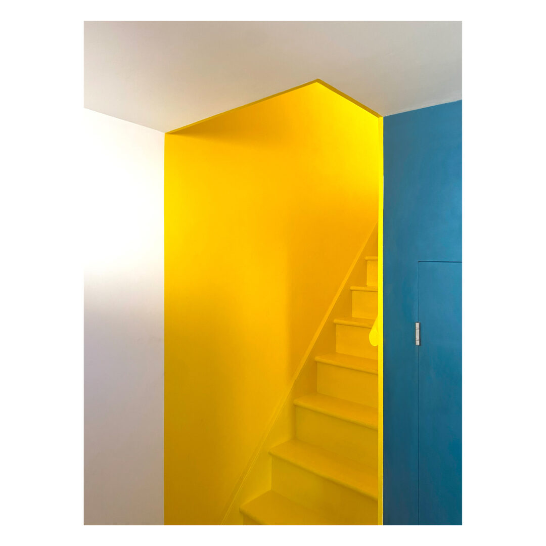 Bright yellow staircase with an enclosed design, nestled between a white wall and a vibrant blue wall with a closed door, creating a bold and colorful architectural contrast.