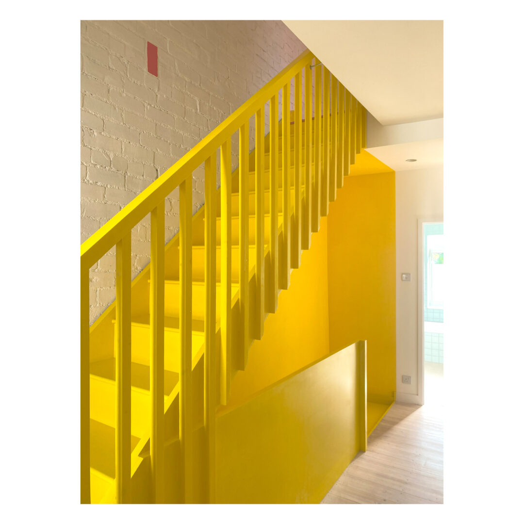 Yellow staircase and railing with wooden steps in a brightly lit interior. A white brick wall is on the left, contrasting with the vibrant yellow. Light wood flooring is visible at the bottom, contributing to the modern and minimalist aesthetic.