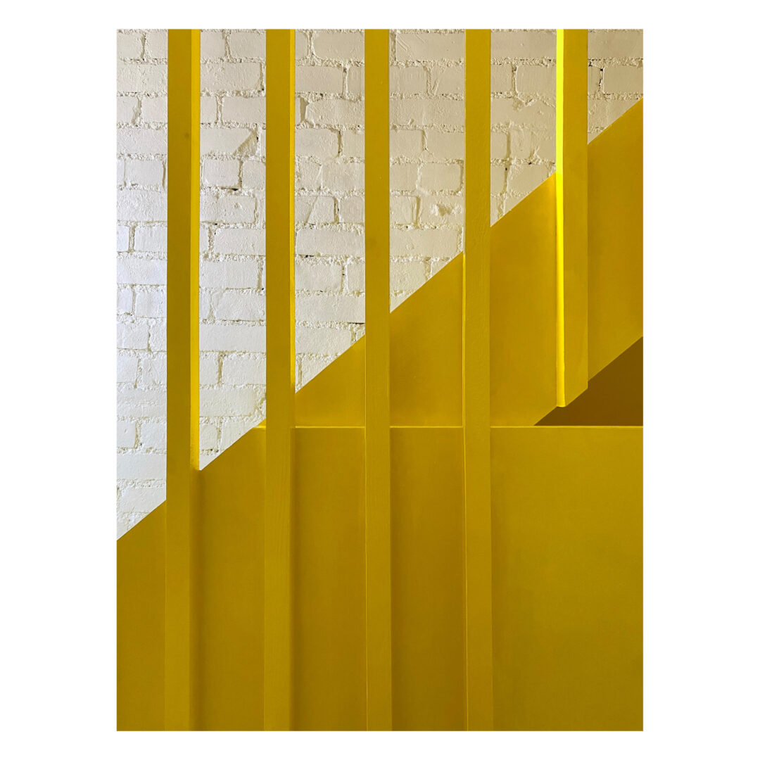Detail of a timber stair balustrade featuring a vertical yellow bars in front of a white brick wall background, creating a dynamic visual contrast.