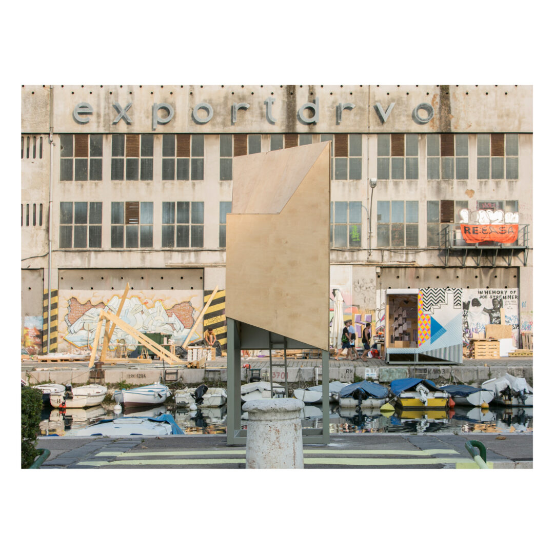 A photograph captures a geometric wooden sculpture crafted by Bristol architects in front of an industrial building with exportdrvo signage. The facade is adorned with colorful murals and graffiti, while several small boats are docked in the calm waterway in the foreground.