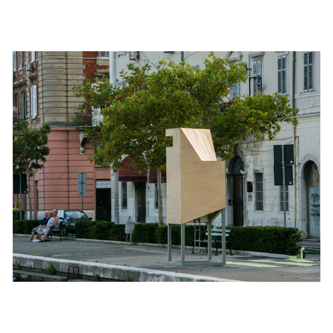 A small, modern wooden structure with a unique angular design stands on a street corner, surrounded by historic buildings. Nearby, a few people sit on a bench under a tree, relaxing. Theres greenery lining the sidewalk.
