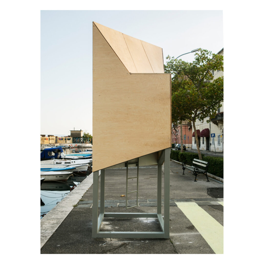 A wooden structure with an angular, modern design stands on a metal frame by a waterfront. The setting includes boats, a few trees, benches, and buildings under a clear sky, creating a peaceful urban scene.