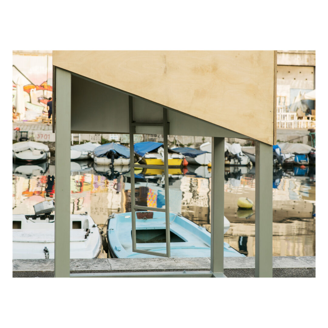 A wooden structure with an open bottom stands in front of a canal filled with docked boats. The water reflects the boats and nearby buildings, creating a mirrored surface. The scene appears calm and quiet.