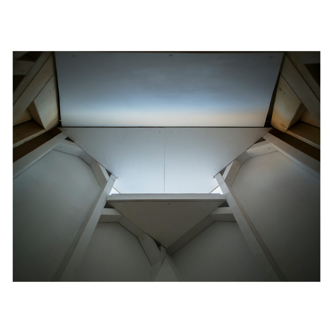 Abstract architectural image with angular lines and geometric shapes. The view looks up towards a narrow skylight, framed by white and gray panels, creating a sense of depth and symmetry. Light enters from above, casting subtle shadows.
