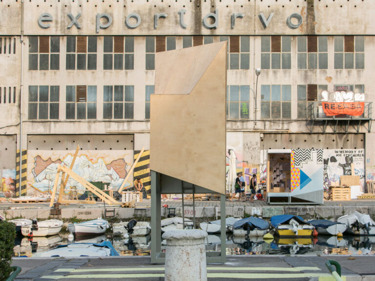 Art installation near a canal with small boats, set in front of a weathered building featuring exportarvo signage. The area boasts vibrant murals and graffiti. A geometric, wood-like structure reminiscent of a house extension in Bristol is centered in the foreground.