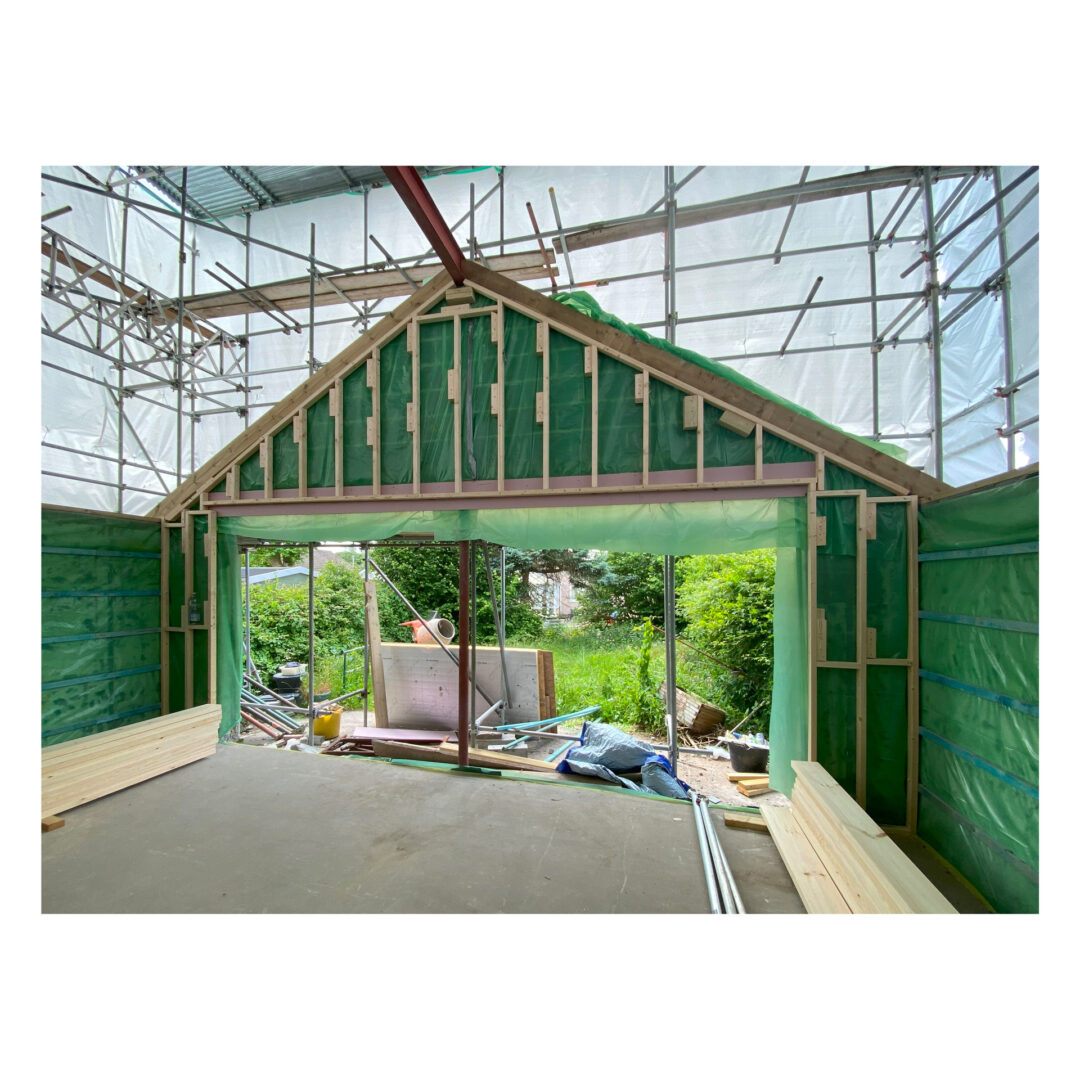 Interior view of a building under construction, featuring a triangular gable end with green insulation. Scaffolding surrounds the structure, and large glass doors open to a garden with trees and greenery outside. Building materials are scattered around.