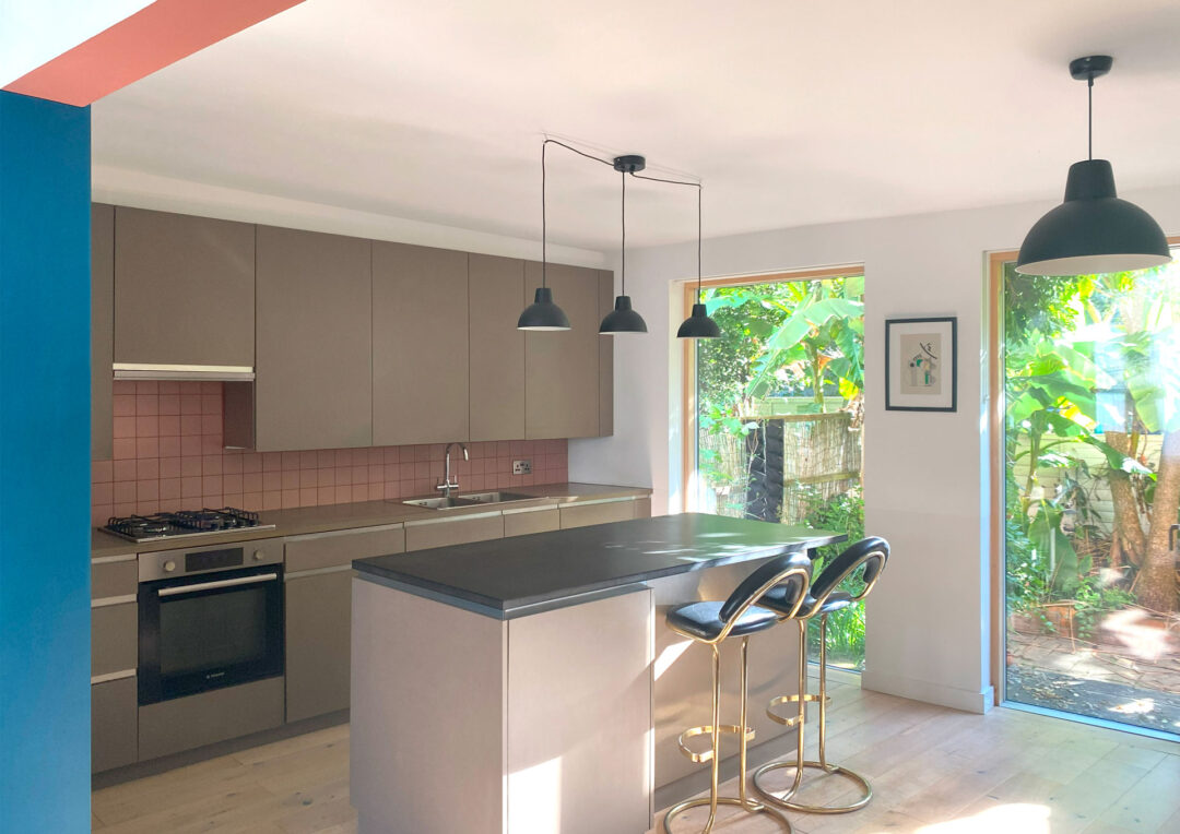 Modern kitchen with beige cabinets, pink backsplash, and a black island with stools. As part of an elegant extension design, three black pendant lights hang above the island. A large window and glass door reveal lush greenery outside, while minimalist decor enhances natural light.