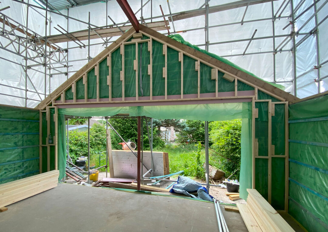 Interior view of a partially constructed building showcases green insulation on walls. Scaffolding surrounds the structure, with lumber and construction materials scattered about. This extension design elegantly transitions to a lush green garden outside, awaiting planning application submission approval.