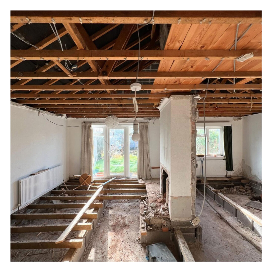 Interior of a house under renovation, with exposed wooden ceiling beams and floor joists. A partially demolished fireplace is visible, and sunlight enters through large patio doors at the back, illuminating the concrete floor and walls.