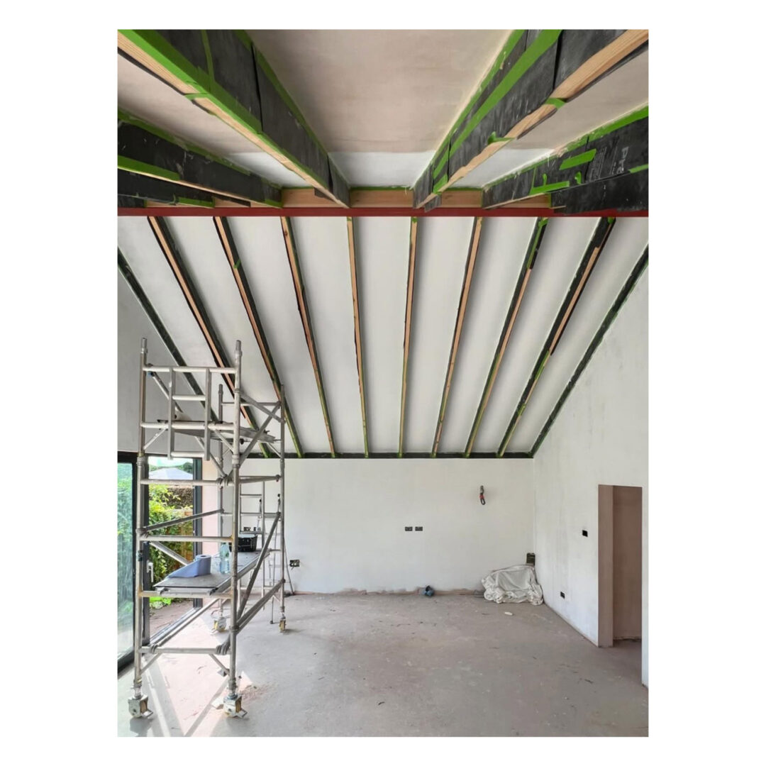 A room under construction with vaulted, partially finished ceilings. Theres a metal scaffold on the left, unfinished walls, and some construction materials on the floor. Natural light enters through a large window on the left.
