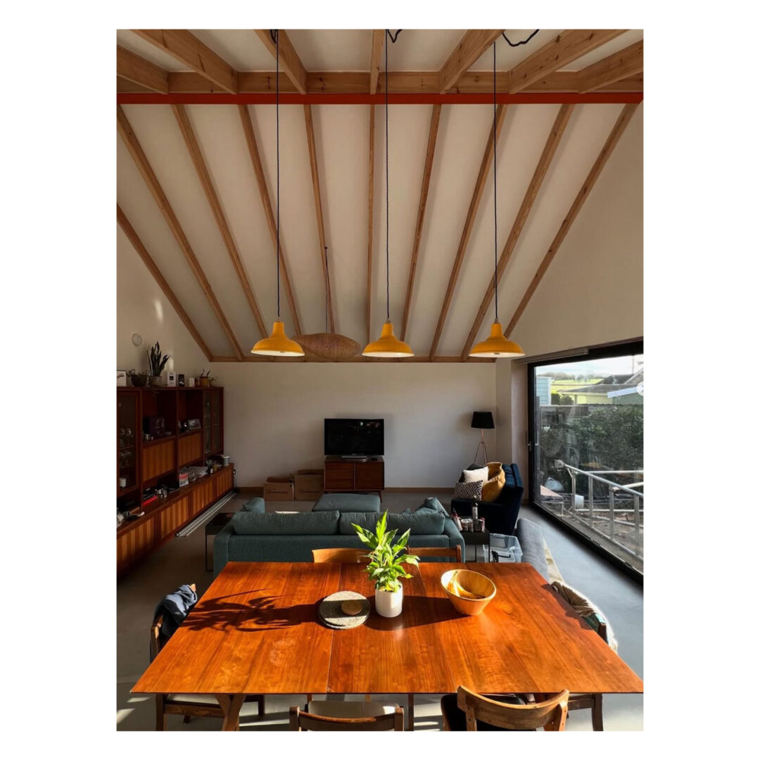 A cozy living room with a wooden dining table in the foreground, adorned with potted plants. Yellow pendant lights hang overhead. In the background, a sofa, TV, and large floor-to-ceiling window are visible, offering a view outside.