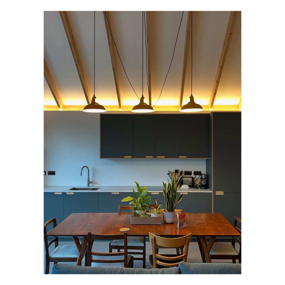 Modern kitchen and dining area with a wooden table, surrounded by chairs. Potted plants on the table. Overhead, three pendant lights hang from a ceiling with exposed wooden beams. A sink, cabinets, and kitchen appliances are visible in the background.